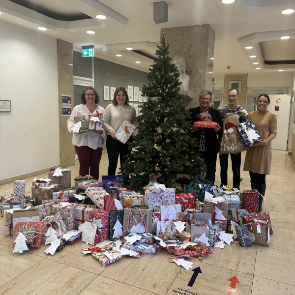 Fünf Personen stehen lächelnd neben einem geschmückten Weihnachtsbaum in einer Bürohalle und halten eingepackte Geschenke in der Hand. Um den Baum herum sind zahlreiche festliche Geschenke auf dem Boden gestapelt.