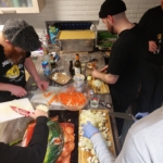 Vier Personen bereiten in einer Küche Essen zu und hacken Gemüse auf Holzbrettern. Verschiedene Zutaten, Messer und Getränke sind auf der Arbeitsplatte verstreut, und im Hintergrund sind eine große Pfanne und Flaschen zu sehen.