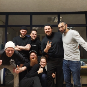 Eine Gruppe von sechs Personen posiert in einem Haus, lächelt und macht spielerische Gesten. Eine Person hält einen Schneebesen, eine andere zeigt ein Friedenszeichen, und andere kauern oder stehen dicht beieinander und zeigen ein Gefühl von Kameradschaft und Spaß.