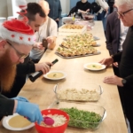 Bei einer Veranstaltung werden Speisen von einem Buffet serviert. Eine Person trägt einen festlichen Hut, eine andere gießt Soße ein, und auf dem Tisch sind mehrere Teller angeordnet. Andere warten mit Tellern, und einige Personen stehen im Hintergrund.