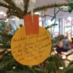 An einem Weihnachtsbaum hängt ein runder gelber Papierschmuck mit einem handgeschriebenen deutschen Text, der den Weltfrieden, keinen Hunger in der Dritten Welt und Hoffnung und Perspektive für alle Betroffenen wünscht. Die Menschen im Hintergrund sind unscharf.