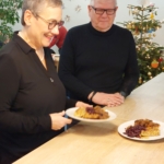 Eine lächelnde Frau serviert an einer Holztheke Essen auf einem Teller, während ein Mann mit weißem Haar und Brille neben ihr steht. Im Hintergrund sind ein geschmückter Weihnachtsbaum und Sternenornamente zu sehen.