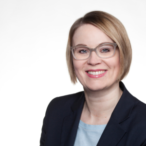 Eine Frau mit kurzen blonden Haaren und Brille lächelt. Sie trägt einen dunklen Blazer über einem hellblauen Oberteil und posiert vor einem schlichten weißen Hintergrund.