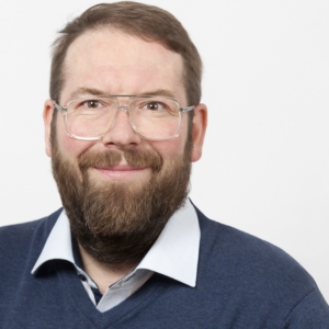 Ein bärtiger Mann mit Brille, der einen blauen Pullover über einem Kragenhemd trägt, lächelt in die Kamera vor einem schlichten weißen Hintergrund.