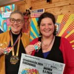 Zwei lächelnde Frauen stehen in einer Halle, tragen Medaillen und halten einen großen Spendenscheck mit der Aufschrift "Jecko & Mio!" für die Feuervogelveranstaltung 2024 in Aachen. Im Hintergrund sind bunte Dekorationen zu sehen.