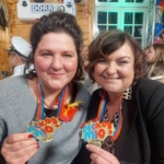 Zwei Frauen, die lächeln und bunte Medaillen mit der Aufschrift "Jecko Mio" in der Hand halten, tragen regenbogenfarbige Bänder. Sie befinden sich auf einer festlichen Indoor-Veranstaltung mit Luftballons und Menschen im Hintergrund.