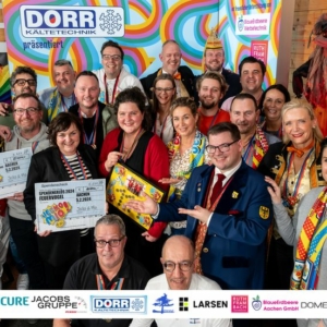 Eine große Gruppe Erwachsener in farbenfrohen Kostümen und mit festlichen Accessoires posiert zusammen in einem Raum, lächelnd und mit Schildern, Auszeichnungen und einem Brettspiel in der Hand. Im Hintergrund sind farbenfrohe Dekorationen und Firmenbanner zu sehen.