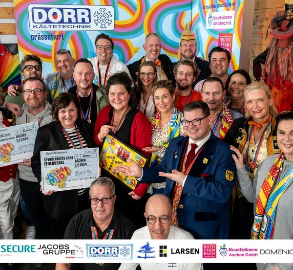 Eine große Gruppe Erwachsener in farbenfrohen Kostümen und mit festlichen Accessoires posiert zusammen in einem Raum, lächelnd und mit Schildern, Auszeichnungen und einem Brettspiel in der Hand. Im Hintergrund sind farbenfrohe Dekorationen und Firmenbanner zu sehen.