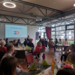 Eine Gruppe von Menschen sitzt an dekorierten Tischen in einem hellen Veranstaltungsraum und hört einem Redner an einem Podium zu. Auf einer Leinwand steht "Suchthilfe Aachen 20 Jahre" mit einem großen roten "20"-Logo.