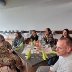 Eine Gruppe von Menschen sitzt um einen mit Getränken, Gläsern und grünen Servietten gedeckten Tisch und beobachtet aufmerksam etwas aus dem Off in einem hellen Raum mit einem Buffet im Hintergrund.