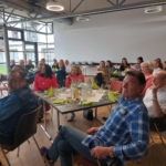 Eine Gruppe von Menschen sitzt in einem hellen Raum mit großen Fenstern um Tische herum und hört aufmerksam jemandem zu, den man nicht sehen kann. Auf den Tischen stehen Getränke, Teller und grüne Servietten.