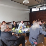 Eine Gruppe von Personen sitzt um einen Tisch in einem modernen Besprechungsraum und hört aufmerksam zu. Auf dem Tisch stehen Getränke, Snacks und Papiernotizen. Weitere Personen sitzen an Tischen im Hintergrund.