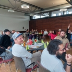 Eine Gruppe Erwachsener sitzt in einem modernen, geräumigen Raum mit Holzfußboden, einige lächeln und sind engagiert, einige heben die Hände. Auf den Tischen stehen Partyhüte und Getränke, und durch große Fenster strömt natürliches Licht herein.