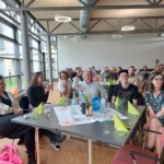 Eine Gruppe von Menschen sitzt um einen Tisch mit Getränken, Snacks und gelben Servietten, lächelt und unterhält sich bei einem Indoor-Event in einem modernen, hellen Raum mit großen Fenstern. Im Hintergrund sitzen weitere Personen.