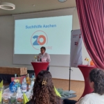 Eine Frau steht auf einem Podium und hält eine Rede für die Suchthilfe Aachen 20 Jahre in einem geschmückten Raum mit einer Leinwand, Tischen mit Getränken und mehreren Zuhörern.