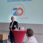Ein Mann im Anzug spricht an einem mit Blumen geschmückten Podium. Hinter ihm steht auf einer Projektion Suchthilfe Aachen und 20 Jahre. Die Gäste haben Platz genommen, auf einem Tisch stehen Flaschen.