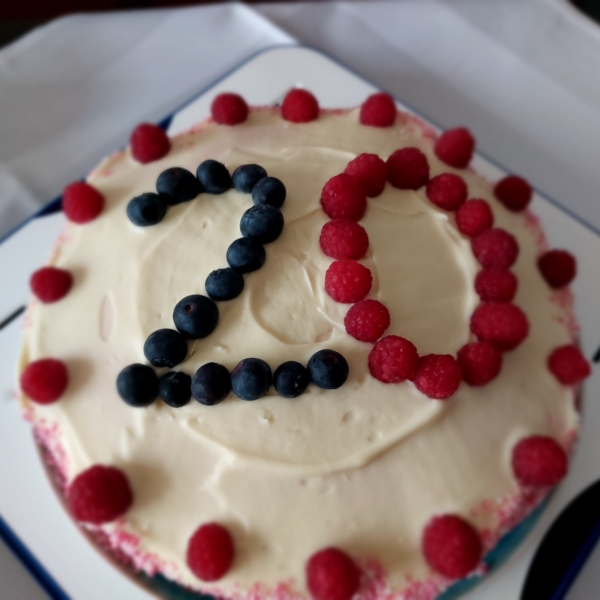 Ein runder Kuchen mit weißem Zuckerguss, der mit Blaubeeren und Himbeeren belegt ist, die die Zahl 20 bilden. Weitere Himbeeren verzieren den Rand der Torte.
