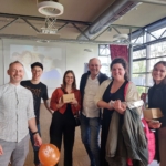 Eine Gruppe von sechs Personen, die lächelnd in einem Innenraum zusammenstehen und kleine Geschenke und einen Luftballon in der Hand halten, mit einem projizierten Bild und Tischen im Hintergrund, was auf ein feierliches oder gesellschaftliches Ereignis hindeutet.