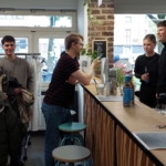 Eine Gruppe junger Erwachsener trifft sich in einem modernen Café. Drei Personen stehen am Tresen und unterhalten sich mit einem Mitarbeiter, während eine weitere Person in der Nähe steht und einen Mantel hält. Auf dem hölzernen Tresen sind Flaschen und Speisekarten zu sehen.