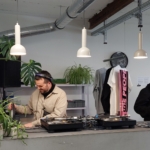 Zwei DJs stehen hinter einem Tresen mit Plattenspielern und Pflanzen. Einer stellt die Regler ein, während der andere lächelt und ein Getränk in der Hand hält. Im Hintergrund hängt Kleidung, darüber sind Hängelampen und freiliegende Rohre zu sehen.