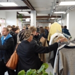 Eine Gruppe von Personen kauft in einer belebten Halle Kleidung ein. Mehrere Personen betrachten die Kleidungsstücke auf den Regalen, während andere durch den Raum gehen. Im Vordergrund ist eine grüne Topfpflanze zu sehen.