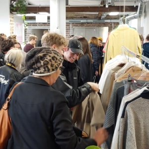 Eine Gruppe von Menschen stöbert in den Kleiderständern einer Markthalle oder eines Secondhand-Ladens. Die meisten sind in Jacken gekleidet; eine Frau trägt einen Hut mit Leopardenmuster und eine braune Tasche. Die Atmosphäre ist geschäftig und lebhaft.