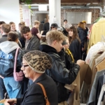 In einem hellen, offenen Geschäft mit freiliegenden Backsteinwänden stöbern viele Menschen in den Kleiderständern. Einige halten Kleidungsstücke hoch, während andere sich unterhalten oder sich durch den Raum bewegen.