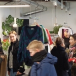 Menschen stöbern in den Kleiderständern eines Secondhand- oder Vintage-Kleiderladens in einer Halle. Pullover und Hemden hängen in der Auslage. Im Hintergrund sind eine Uhr und eine Topfpflanze zu sehen. Die Atmosphäre ist geschäftig und gesellig.