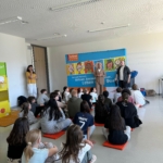 Eine Gruppe von Kindern sitzt auf orangefarbenen Matten und blickt auf drei Erwachsene, die in einem Klassenzimmer vor einer bunten Präsentationstafel stehen. Auf der Tafel sind Cartoon-Gesichter und ein deutscher Text über eine gesunde Lebensweise zu sehen.