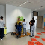 In einem hellen Raum stehen mehrere Kinder um ein interaktives Exponat herum und beschäftigen sich mit der Ausstellung. Orangefarbene Matten mit Illustrationen liegen auf dem Boden verstreut, und zwei Erwachsene beaufsichtigen sie.