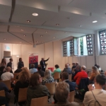 Eine Gruppe von Personen sitzt in einem hellen Konferenzraum und hört aufmerksam einem Redner zu, der nach vorne gestikuliert. Drei Personen stehen in der Nähe, und an der Wand hinter ihnen sind bunte Plakate zu sehen.
