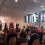 In einem hellen Raum mit großen Fenstern und dekorativen Fenstergittern sitzt eine Gruppe von Menschen vor einem Podium von Rednern. Das Publikum hört der Diskussion aufmerksam zu.
