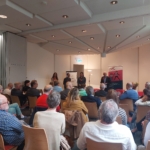 Eine Gruppe von Personen sitzt vor einer Gruppe von vier Rednern, die vorne in einem gut beleuchteten Konferenzraum stehen und hinter denen Banner und Poster angebracht sind.