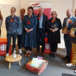 Acht Erwachsene stehen auf einer Bühne, einige halten Bücher in der Hand, alle tragen orangefarbene herzförmige Anstecknadeln. Ein Korb mit Büchern steht vor ihnen auf dem Boden. Hinter ihnen sind Banner mit Logos und Texten angebracht.