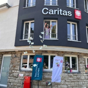 Eine Person steht an einem offenen Fenster eines Caritas-Gebäudes und wirft Euro-Banknoten hinaus. Das Geld ist in der Luft über einem Bürgersteig zu sehen, in dessen Nähe sich ein Caritas-Schild, eine Fahne und eine Spendenbox befinden.