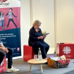 Eine Frau sitzt auf einem roten Würfelsessel und liest auf der Bühne aus einer Zeitung vor. Neben ihr stehen ein mit Büchern und Geschenken gefüllter Korb, eine Flasche Wasser und ein Glas auf einem kleinen Tisch. Hinter ihr ist ein Banner mit Kindern und einem Text in deutscher Sprache zu sehen.