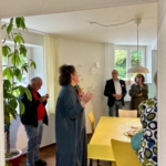 Eine Gruppe von Menschen steht in einem hellen Raum mit einem gelben Tisch, spricht und isst. Eine Frau in einem blauen Kleid gestikuliert, während sie spricht, während die anderen zuhören. Der Raum hat große Fenster und grüne Pflanzen.