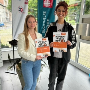 Zwei Frauen stehen in einem Innenraum, lächeln und halten Urkunden für einen Hip-Hop-Wettbewerb 2023 in der Hand. Hinter ihnen hängen Banner mit "Suchthilfe Aachen" und einem Logo, links davon ist eine Pflanze zu sehen.