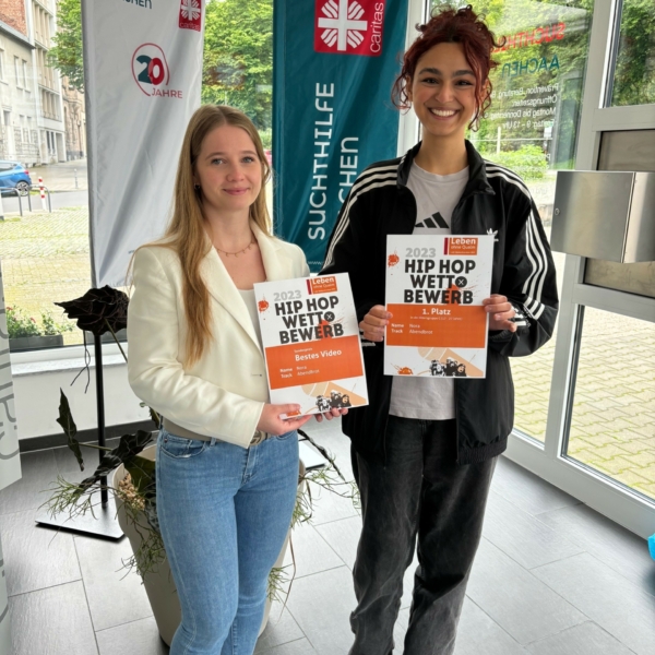 Zwei Frauen stehen in einem Innenraum, lächeln und halten Urkunden für einen Hip-Hop-Wettbewerb 2023 in der Hand. Hinter ihnen hängen Banner mit "Suchthilfe Aachen" und einem Logo, links davon ist eine Pflanze zu sehen.