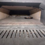 Im hinteren Teil eines großen, leeren Holzofens mit Kachelboden und schwarzen, rußigen Wänden steht eine Metallpfanne mit Essen. Die Ofentür ist geöffnet und gibt den Blick auf das Innere und eine vergitterte Metallschwelle frei.