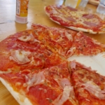 Eine Nahaufnahme einer aufgeschnittenen Pizza mit Schinken und Käse auf einem Pappteller, der auf einem Holztisch steht. Im Hintergrund stehen weitere Pizzateller und einige Flaschen mit Getränken.