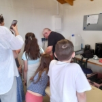 Eine Gruppe von Kindern und Erwachsenen versammelt sich um einen Mann, der auf einem Tisch in einem Raum etwas vorführt. Eine Person macht ein Foto mit einem Telefon. Im Hintergrund sind Küchengeräte zu sehen.