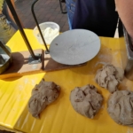 Vier Teigstücke liegen auf einem gelben Tisch, daneben eine Metallpresse, eine runde weiße Platte, ein Behälter mit Mehl und eine Person, die den Teig in einem Metalleimer rührt. Eine weitere Person steht in der Nähe.