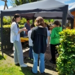 Eine Gruppe von Menschen, darunter Kinder und Erwachsene, steht an einem sonnigen Tag unter einem grauen Vordach in einem Garten. Eine Frau in Weiß gestikuliert, während die anderen zuhören. Im Hintergrund sind Grünflächen und blauer Himmel zu sehen.