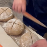 Auf einem Küchentisch ritzt eine Person mit einem Messer drei runde, bemehlte Brotlaibe vor dem Backen mit Kreuzmustern ein.