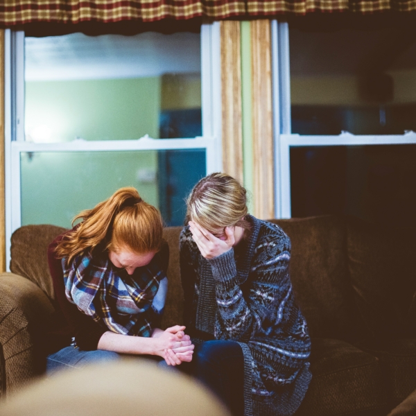 Zwei Personen sitzen eng beieinander auf einer Couch in einem Wohnzimmer, beide mit gesenktem Kopf nach vorne gelehnt, eine davon mit einer Hand, die ihr Gesicht bedeckt, was darauf hindeutet, dass sie einen Moment des Trostes oder der Trauer miteinander teilen.