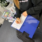Eine Person, die auf einem schwarzen Stuhl sitzt, füllt ein Formular auf einem Klemmbrett mit blauem Umschlag aus. In der Nähe sind Zeitschriften auf einem Tisch gestapelt, und im Hintergrund ist ein Teil einer Pflanze zu sehen.