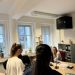Zwei Frauen stehen im Gespräch mit einer Frau, die an einem Schreibtisch in einem Büro mit großen Fenstern, Pflanzen, Regalen und Papieren an den Wänden sitzt. Die Szene scheint eine Besprechung oder Beratung zu sein.