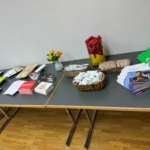 Ein Schautisch mit Stapeln von Broschüren, Zeitschriften und Postkarten, Körbe mit eingepackten Snacks, ein Glas mit roten Servietten, eine kleine Vase mit Blumen und ein Behälter mit bunten Rührstäbchen auf einem Holzboden.