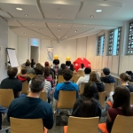 Eine Gruppe von Menschen sitzt auf Stühlen vor einer kleinen Bühne, auf der drei Darsteller stehen, zwei mit gelben Masken und einer mit einem roten Schirm. Der Raum ist hell mit weißen Wänden und großen Fenstern.